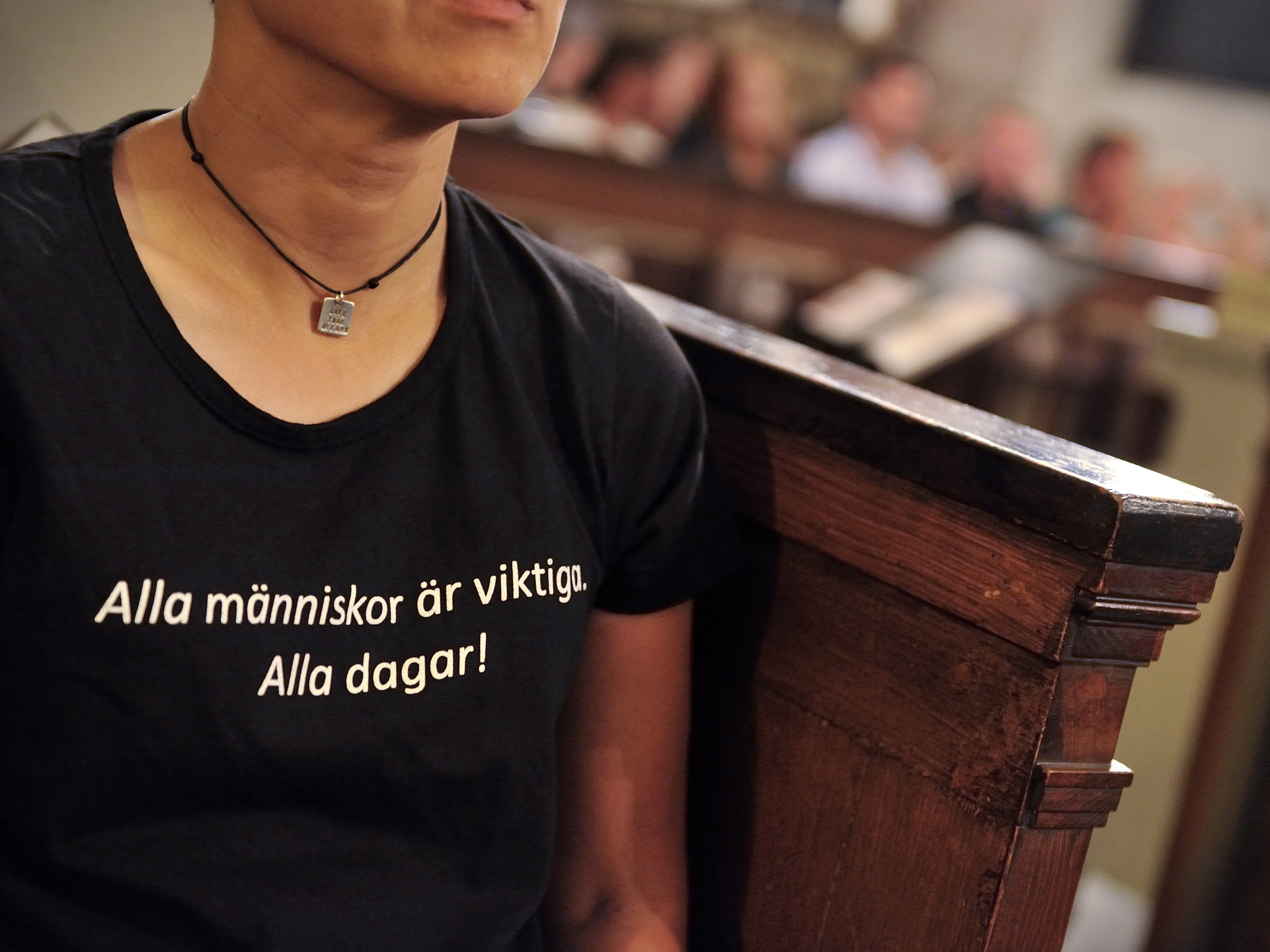 En person i svart t-shirt med texten Alla människor är viktiga alla dagar, sitter i en kyrkbänk.