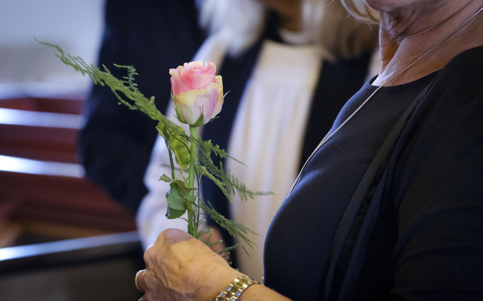 En kvinna i svarta kläder håller en ljusrosa ros. Kyrkbänkar i bakgrunden.