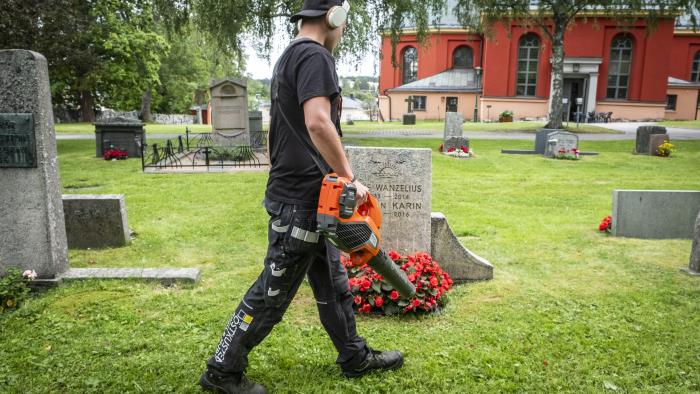 En ung kille går med en lövblås runt bland gravstenarna på en kyrkogård.