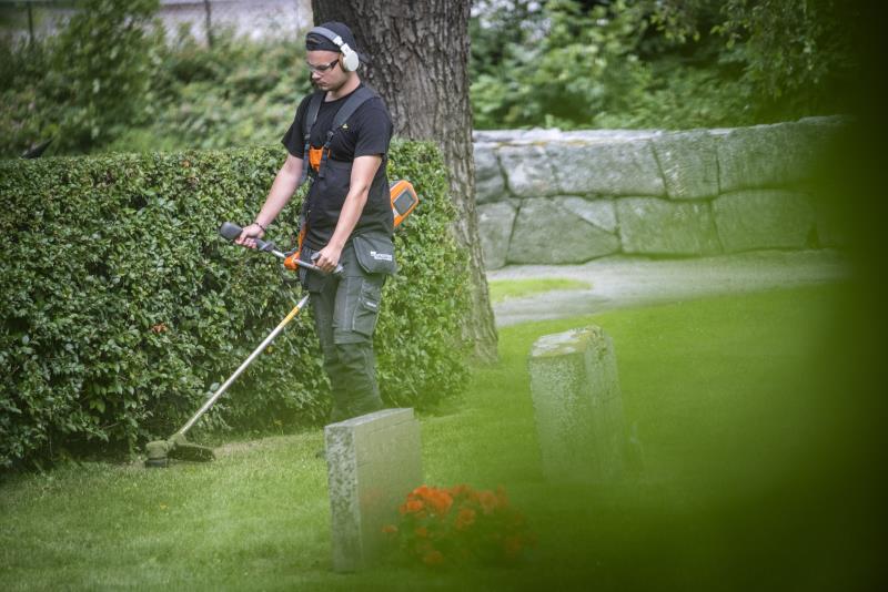 En ung kille klipper gräset runt en häck med en trimmer på en kyrkogård.