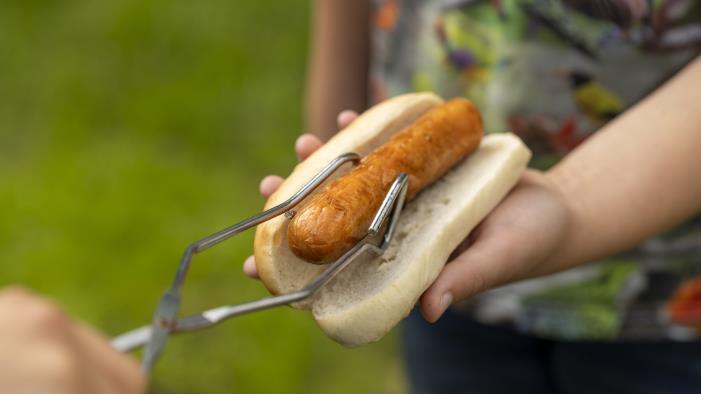 Någon lägger med tång en grillad korv till en person som håller fram ett korvbröd.