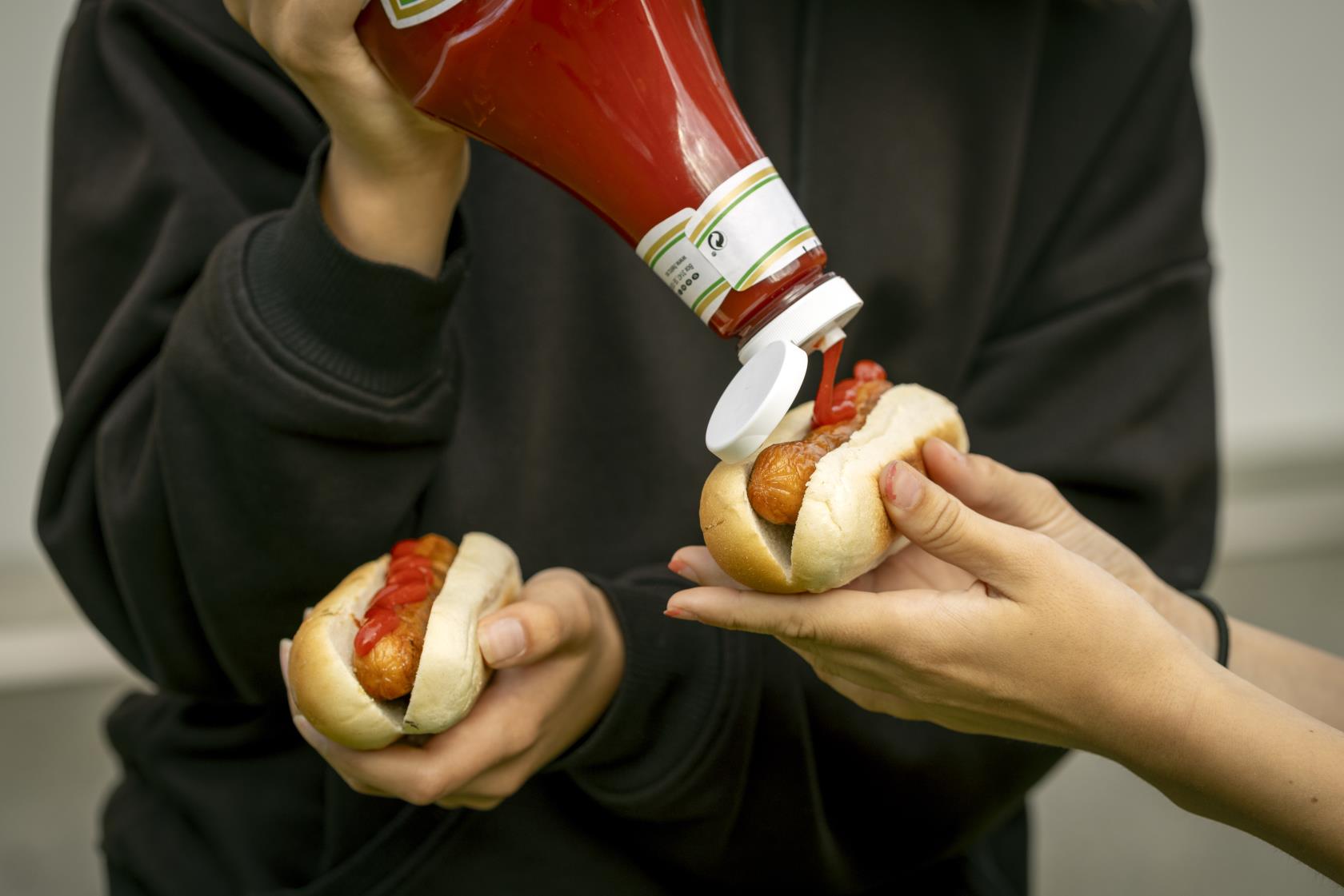 En person sprutar ketchup på en varmkorv som någon håller fram.