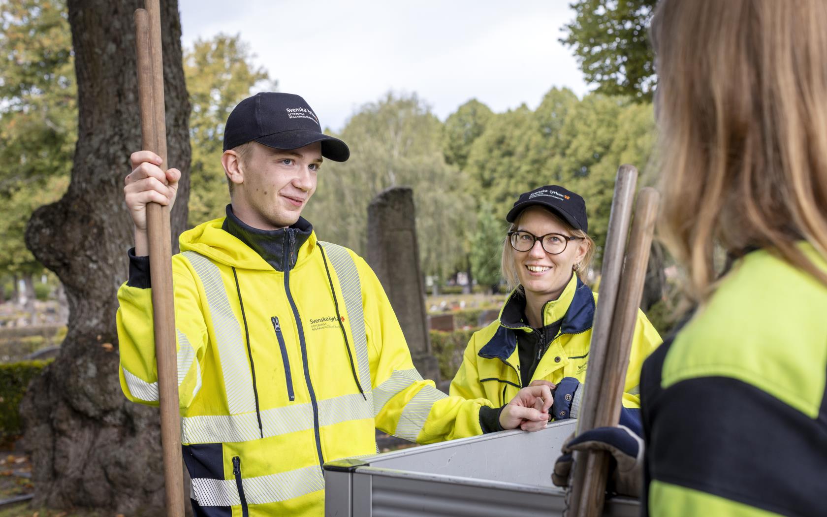 Några kyrkogårdsarbetare i gula varselkläder står och samtalar.