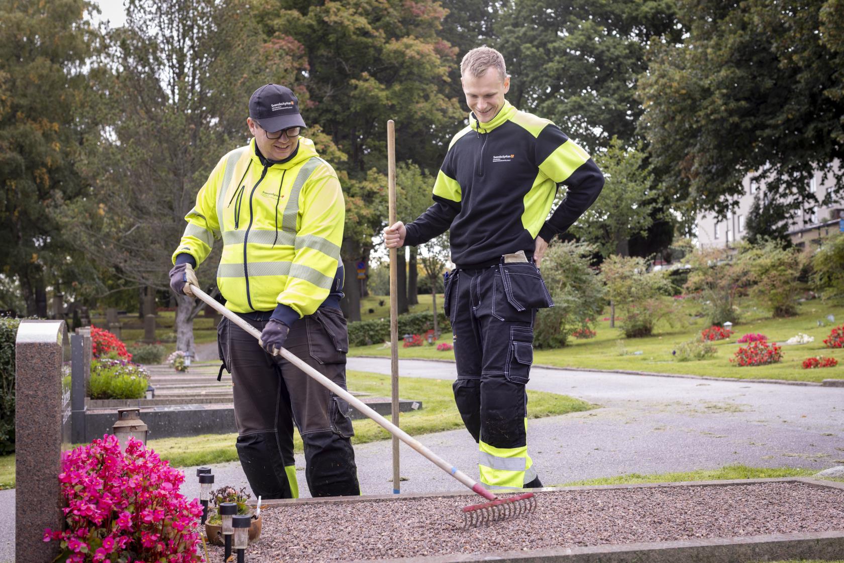 Två kyrkogårdsarbetare i gula varselkläder krattar gruset på en grav.