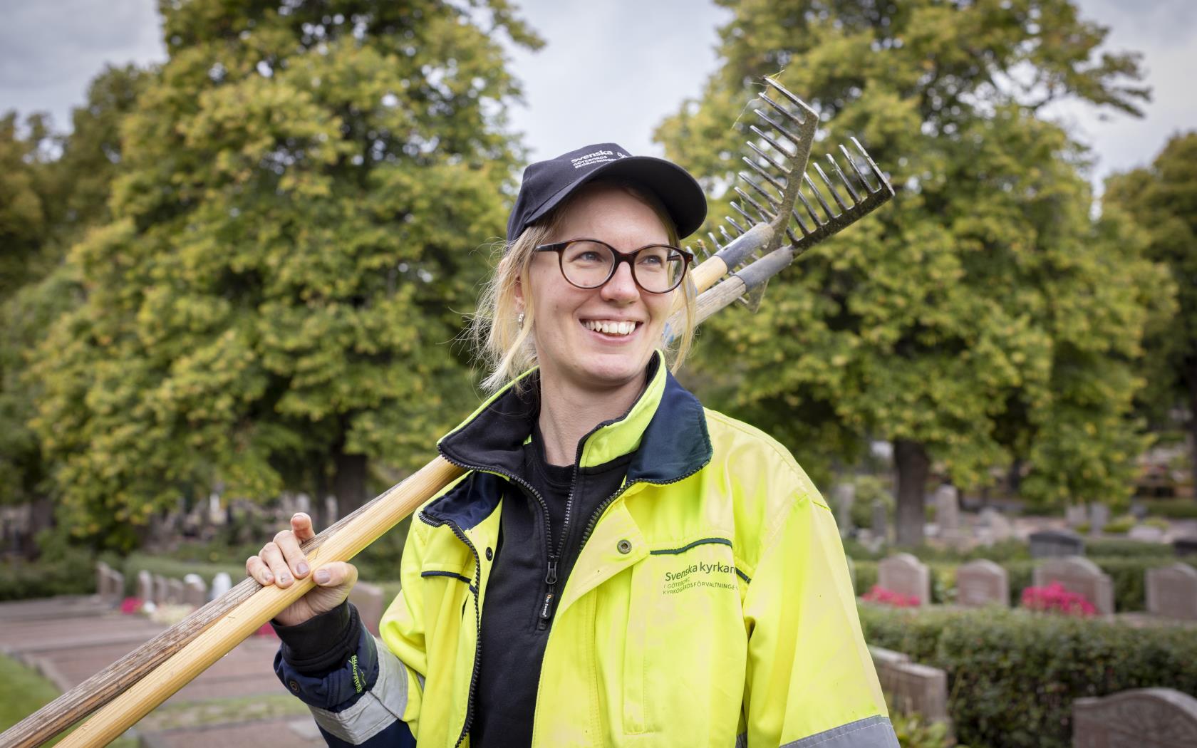 En kvinna i gula varselkläder och keps står med två krattor över axeln på en kyrkogård. Hon ler stort.