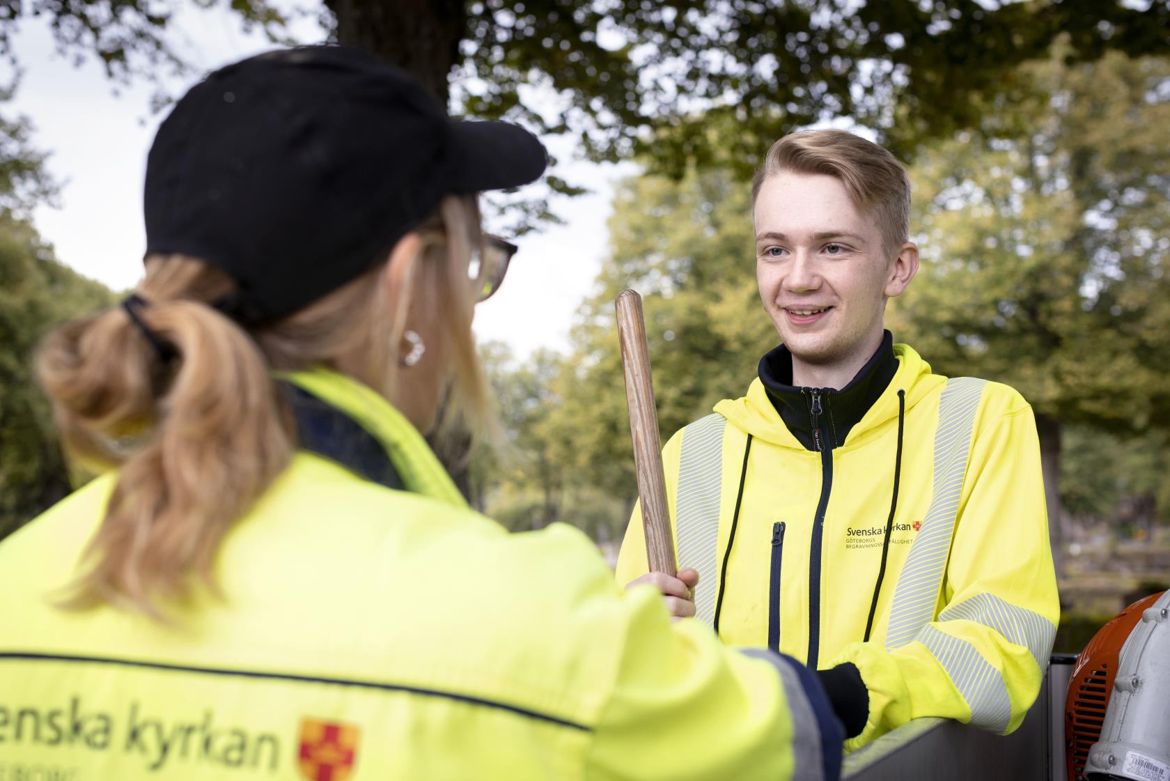 Två kyrkogårdsarbetare i gula varselkläder står och samtalar.