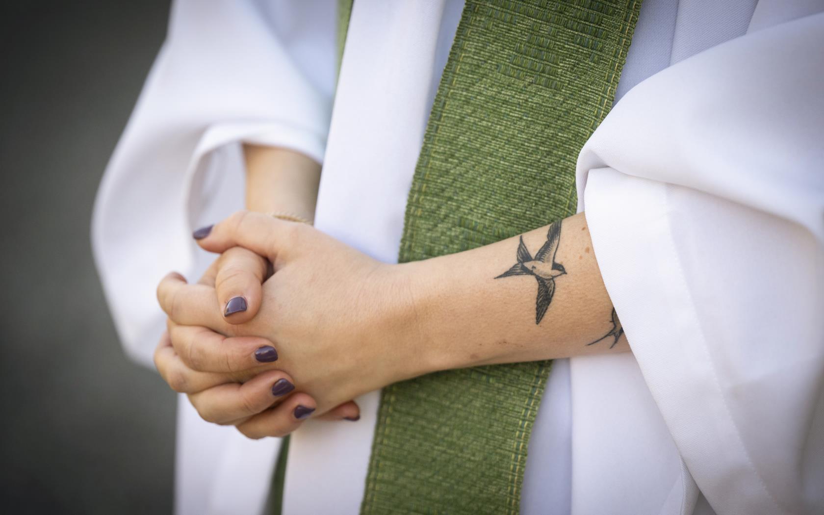 En präst med grön stola, lila nagellack och tatueringar står med knäppta händer.