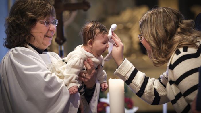 En kvinnlig präst håller en bebis som precis döpts i famnen och mamman torkar barnets huvud.