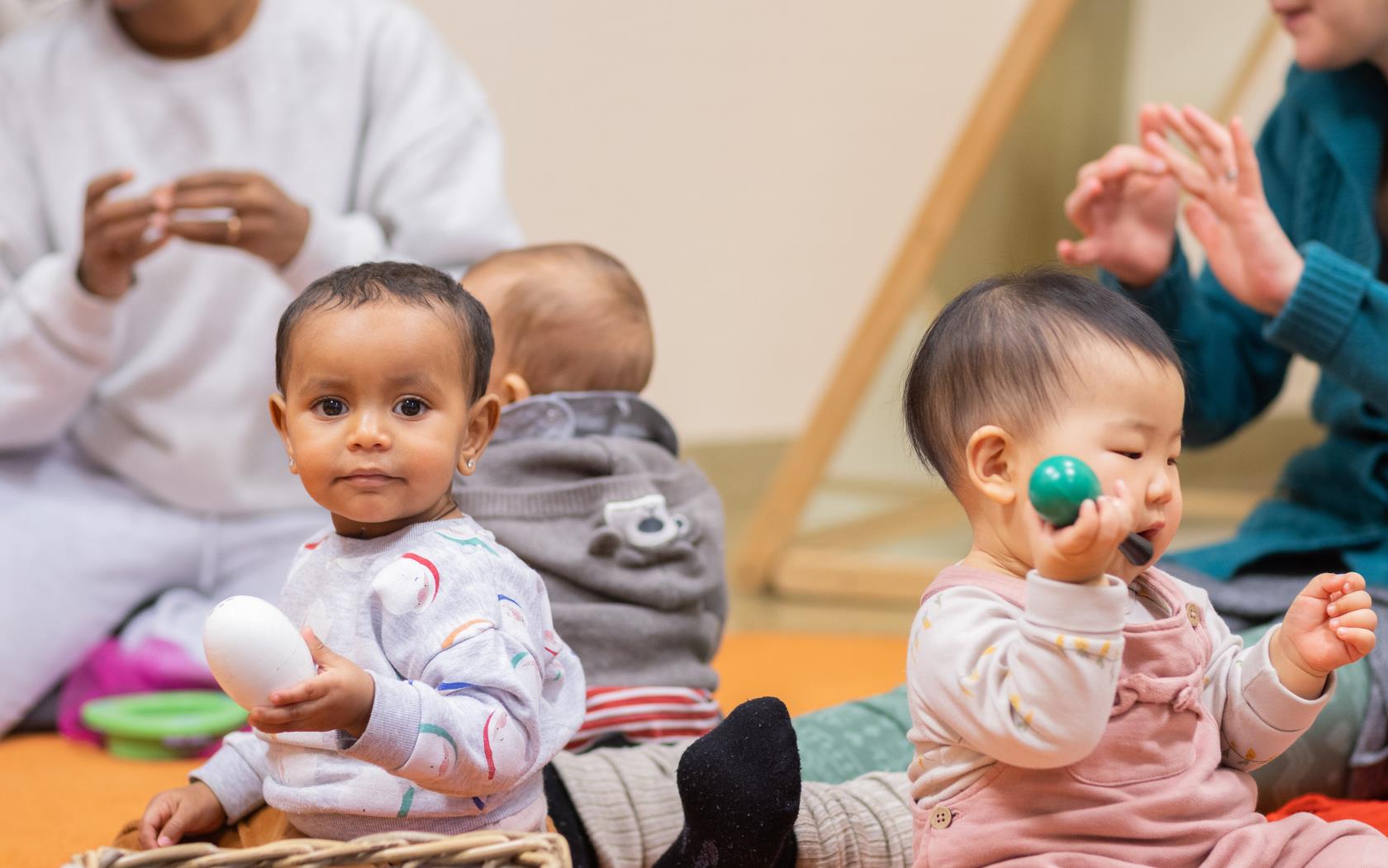 Tre bebisar sitter på golvet. Runt om sitter deras mammor och sjunger.