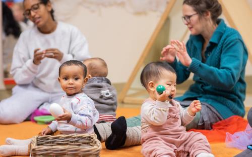 Tre bebisar sitter på golvet. Runt om sitter deras mammor och sjunger.