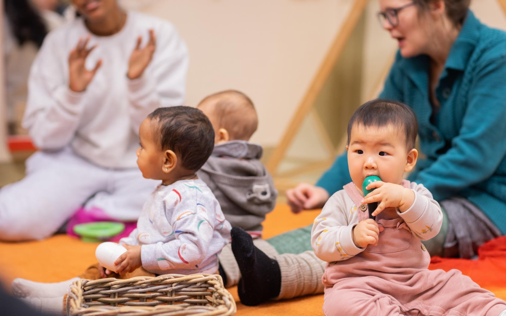 Tre bebisar sitter på en matta. Deras föräldrar sitter runt om.