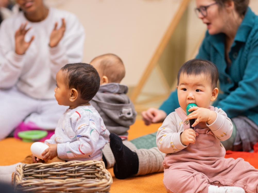 Tre bebisar sitter på en matta. Deras föräldrar sitter runt om.