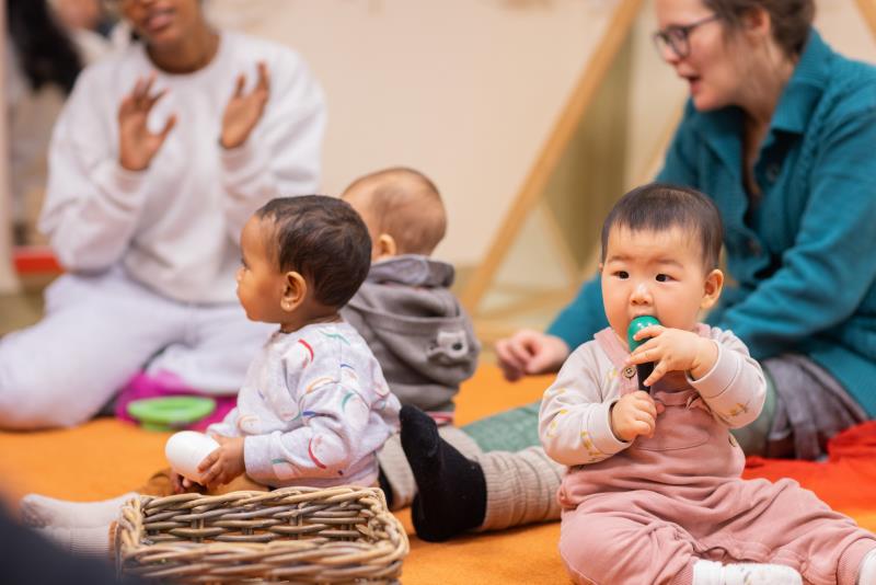 Tre bebisar sitter på en matta. Deras föräldrar sitter runt om.