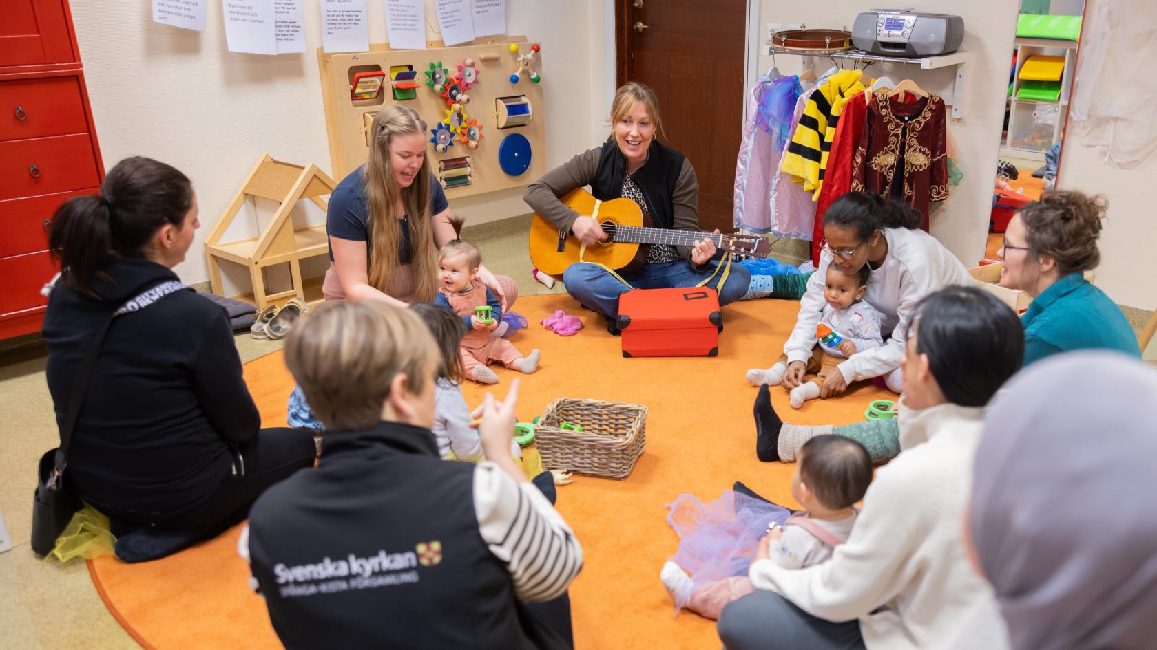 På en rund matta sitter några mammor i en ring, några bebisar innanför och en kvinna med gitarr.