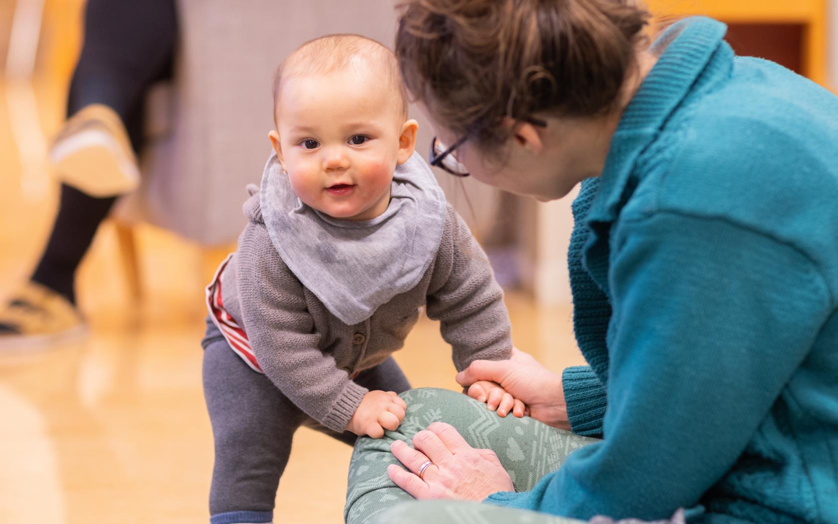 En bebis ställer sig trevande upp, stödd mot sin mamma.