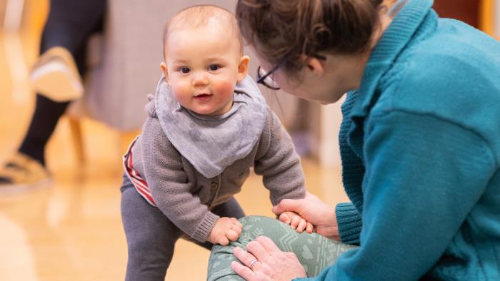 En bebis ställer sig trevande upp, stödd mot sin mamma.