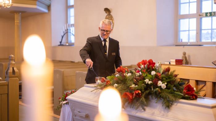 En kyrkvaktmästare tänder ett ljus på en vit kista som har en krans av amaryllis, tallkvistar och vita rosor.
