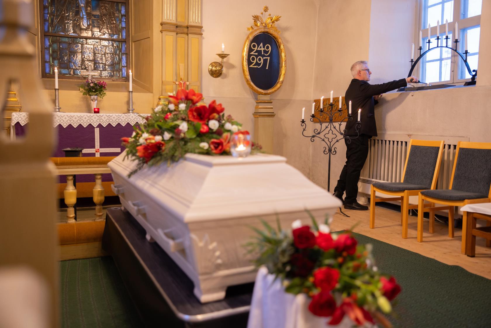 En kyrkvaktmästare tänder ljus i ett fönster. I koret står en vit kista med röda och vita blommor på.