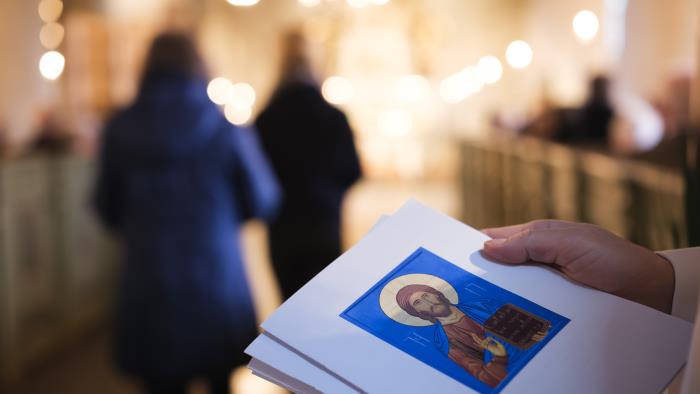 I förgrunden en hand som håller i en agenda. I bakgrunden ser vi ett upplyst kor och människor som rör sig mot det.