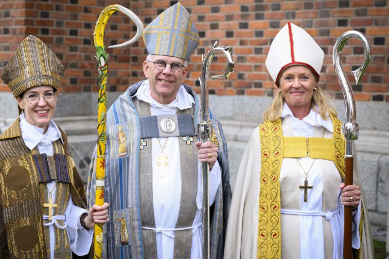 Ärkebiskop står framför en tegelkyrka med två kvinnliga biskopar på var sin sida. Alla bär liturgiska kläder och biskopsstav.