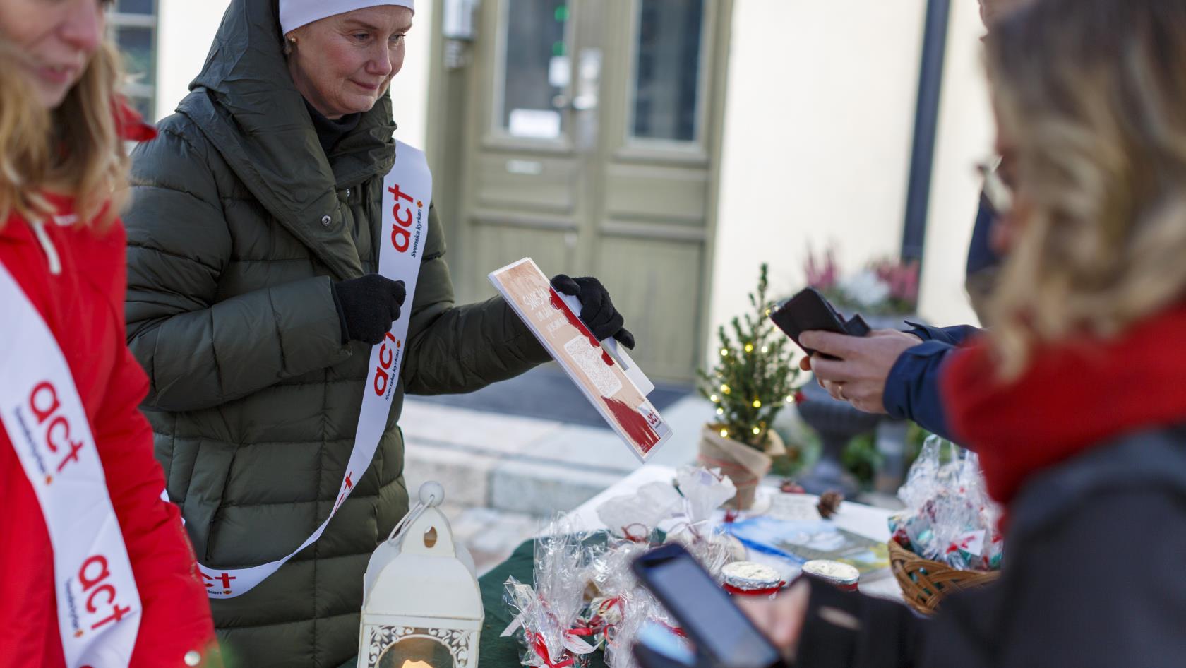 Två kvinnor med Act Svenska kyrkan banderoller över axeln säljer lotter och kakor på julmarknad.