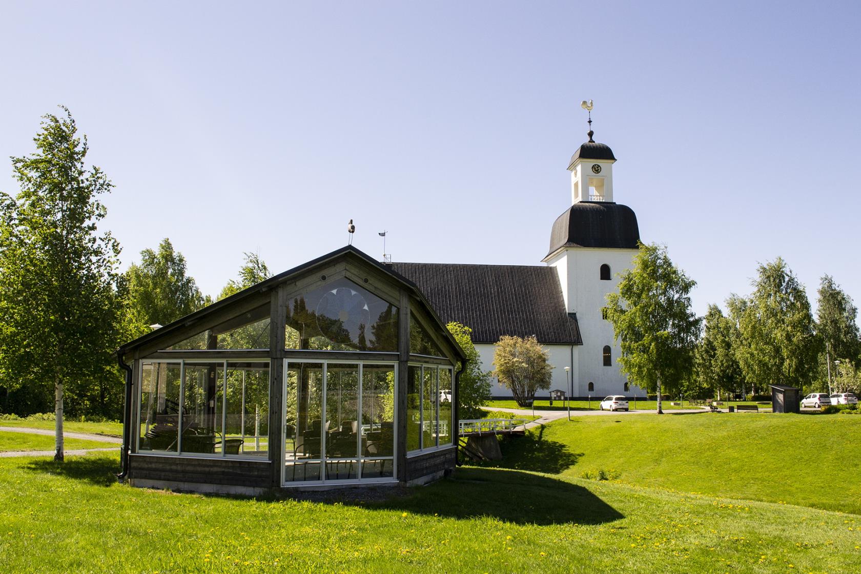 Bilden visar i förgrunden växthuset vid församlingshemmet i Kusmark. I bakgrunden ser vi Kusmarks kyrka. 