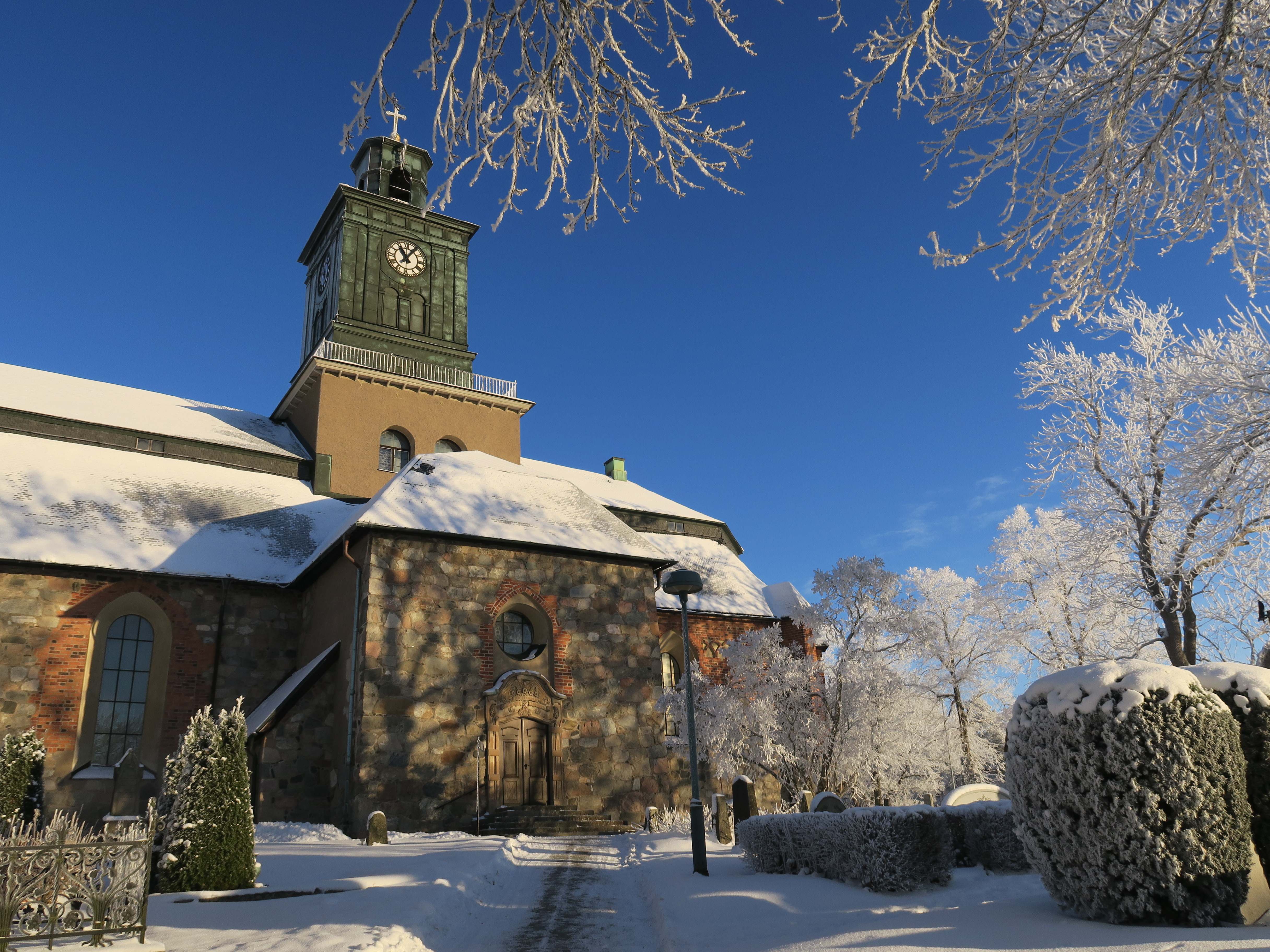 Vårfrukyrkan, en gammal korskyrka från 1100-talet med torn, sträcker sig mot en blå vinterhimmel. Runt omkring syns snötäckta träd.