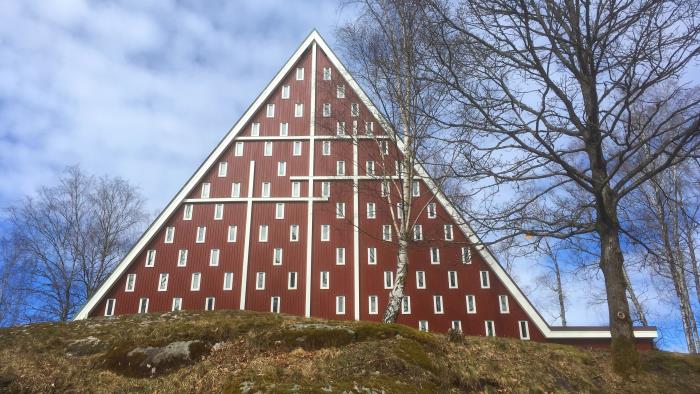 Röd träkyrka med tre vita kors på gaveln. Omgiven av träd utan löv.