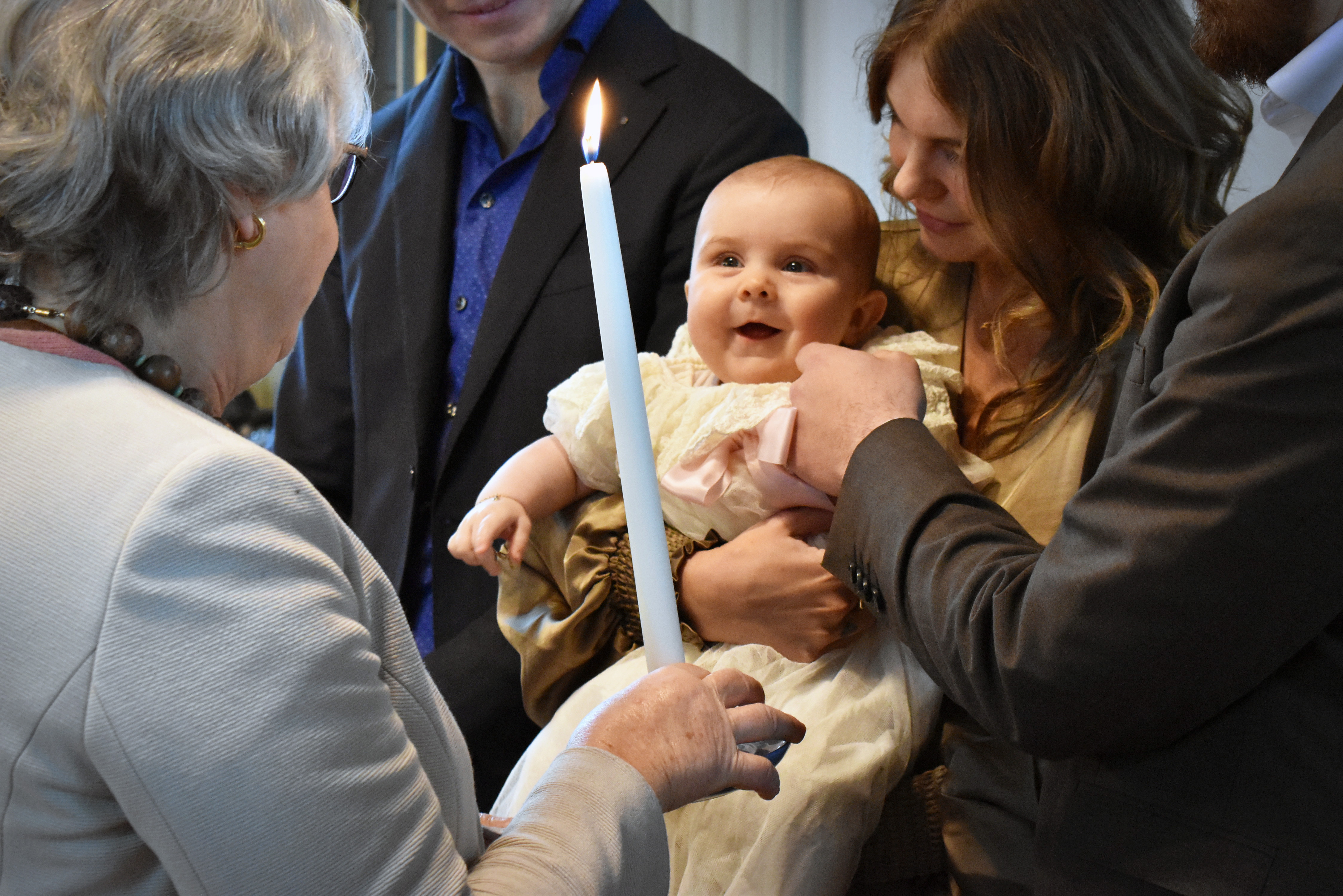 Litet barn i mammas famn tittar glatt på ett tänt stearinljus som äldre kvinna håller fram.