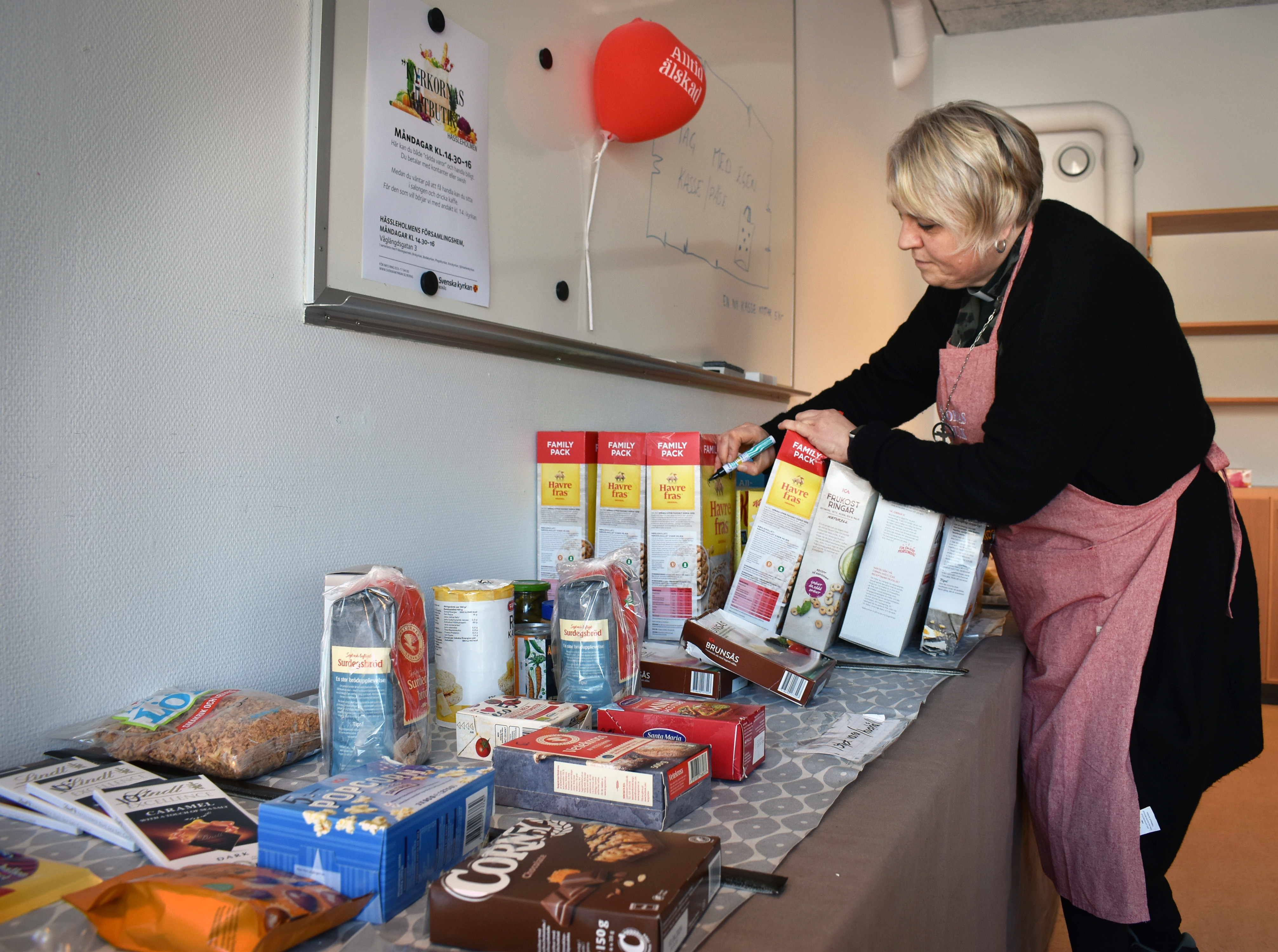 Kvinna i butiksförkläde med penna i handen och matvaror framför sig.