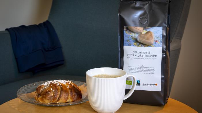 En kaffekopp, en kanelbulle och en påse med kaffe från Svenska kyrkan står på ett bord.