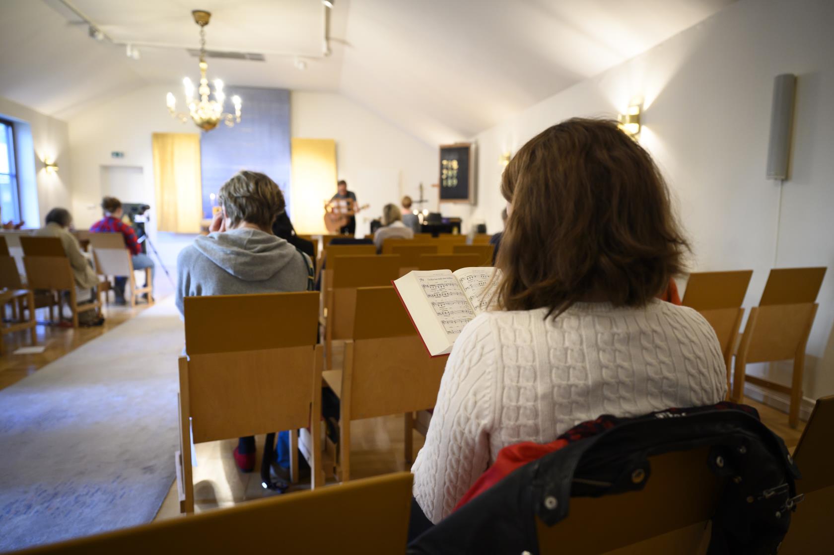 En kvinna sitter i en kyrkbänk med en psalmbok i handen. Flera besökare sitter på raderna framför.