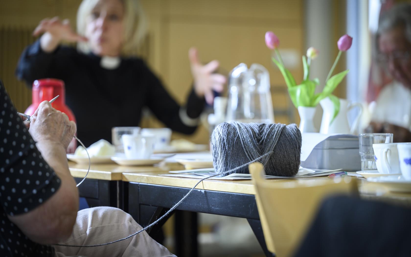 Någon sitter vid ett bord och stickar. Vid bordet syns en kvinnlig präst gestikulera med armarna.