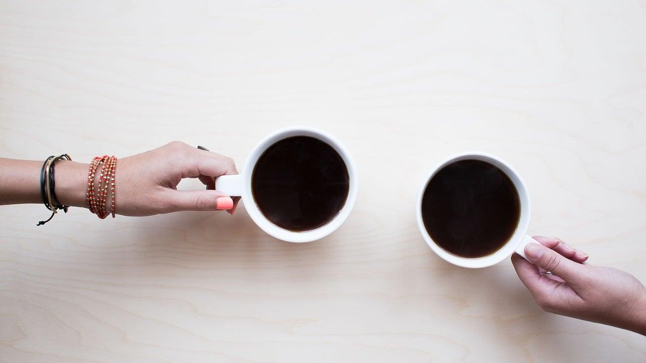 Två händer håller i varsin kopp med kaffe som står på ett bord.