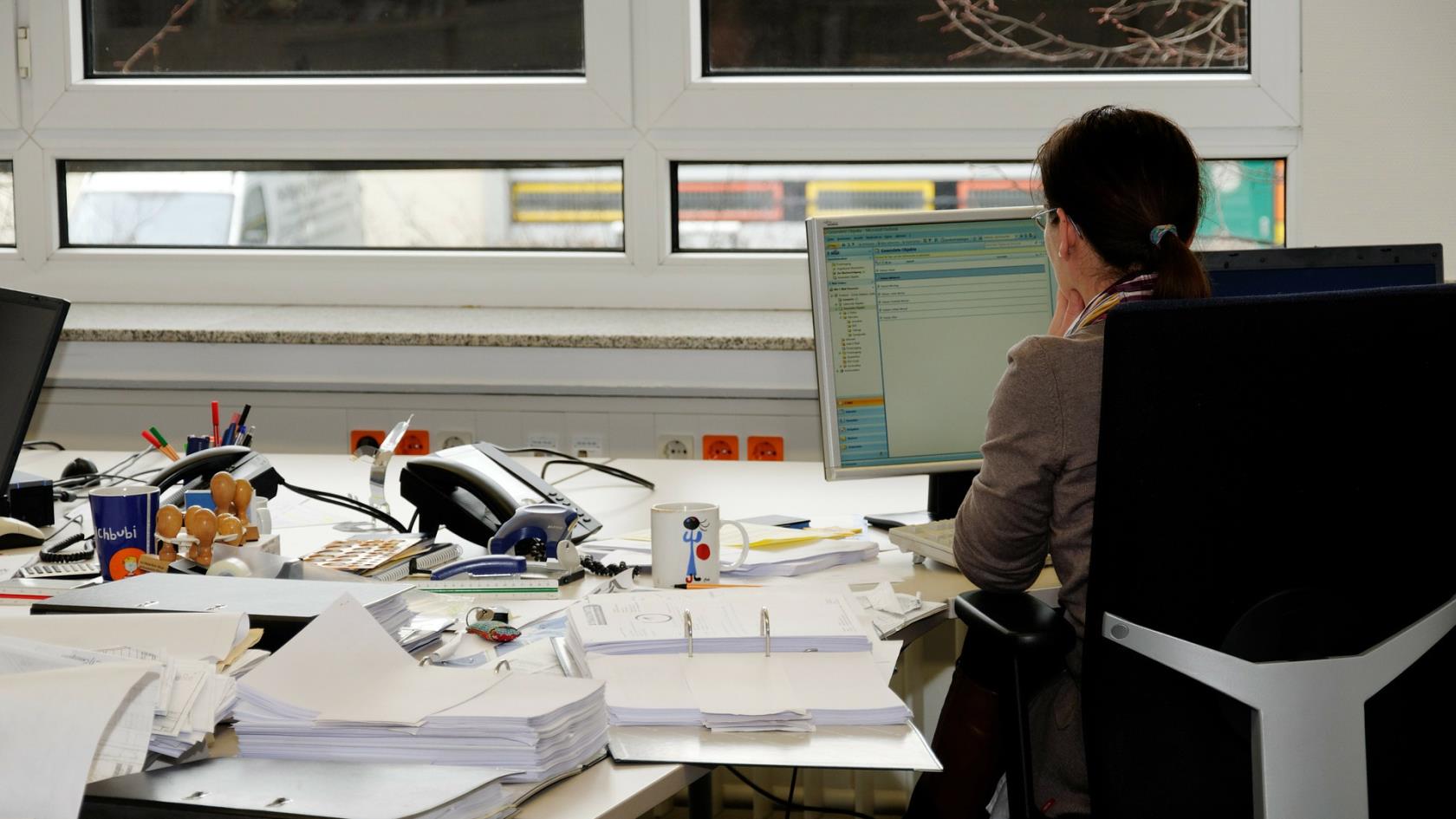 En kvinna sitter på en kontorsstol vid ett stort bord fyllt med papper, en telefon och en dator