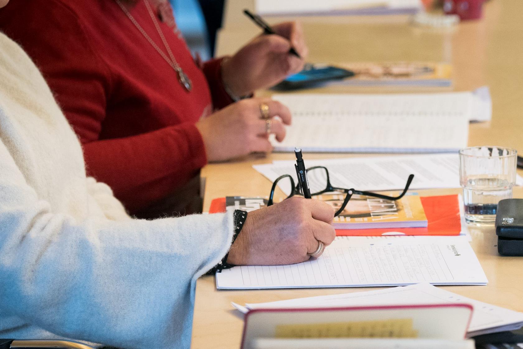 Två personer sitter på möte. På ett bord framför dem finns pennor, papper, broschyrer, glasögon, vattenglas.