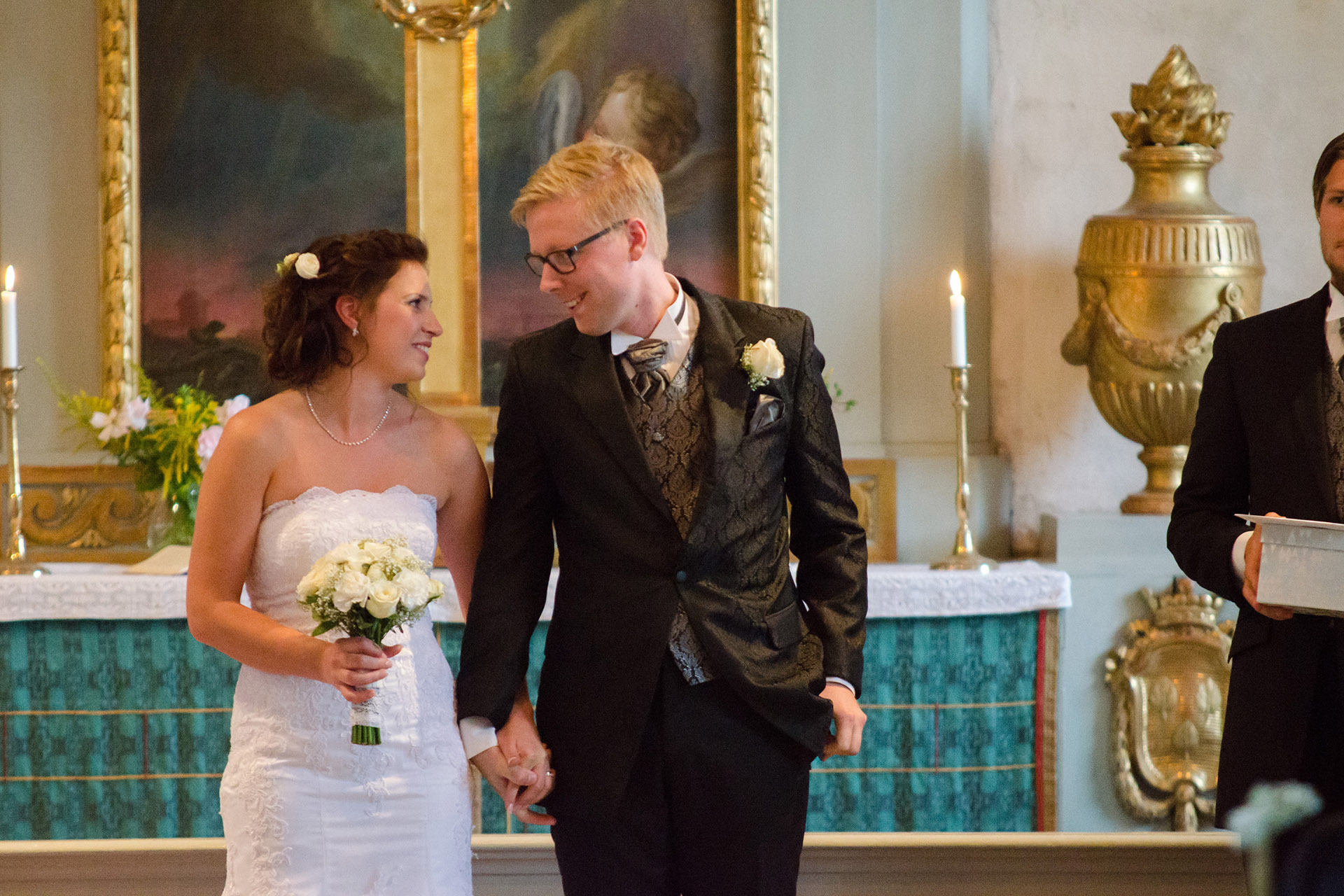 Vigselparet, en kvinna och en man i bröllopskläder, står med altaret bakom sig. De ser på varandra.