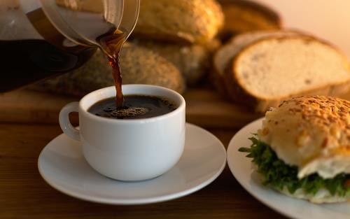 Person häller upp kaffe i en kopp och bredvid finns en bulle och bröd.