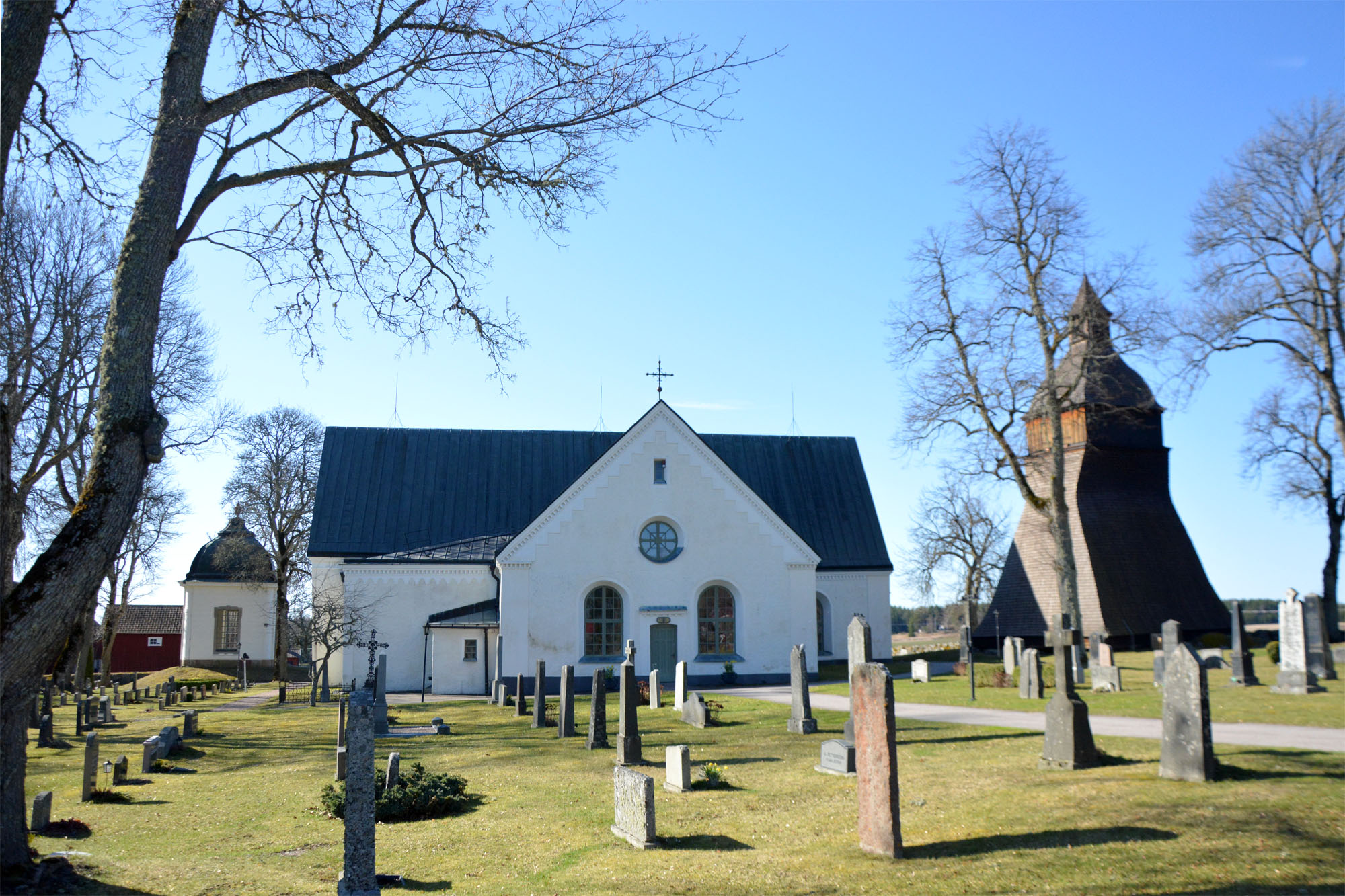 Vit stenkyrka med tjärad klockstapel till höger och litet gravkor till vänster samt gravstenar och träd i förgrunden.