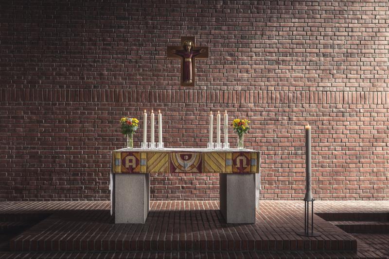 Ett altare med blomvaser och tända ljus står i ett rum med tegelgolv och tegelväggar. Ett krucifix hänger på väggen.