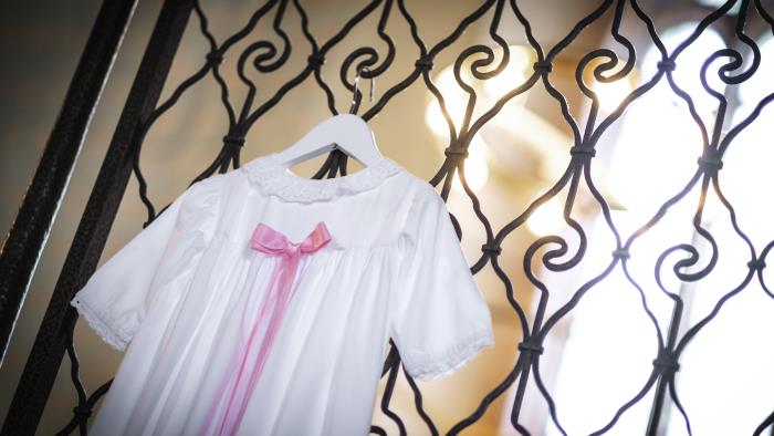 En vit dopklänning med rosa rosett hänger på en metallgrind i kyrkan.