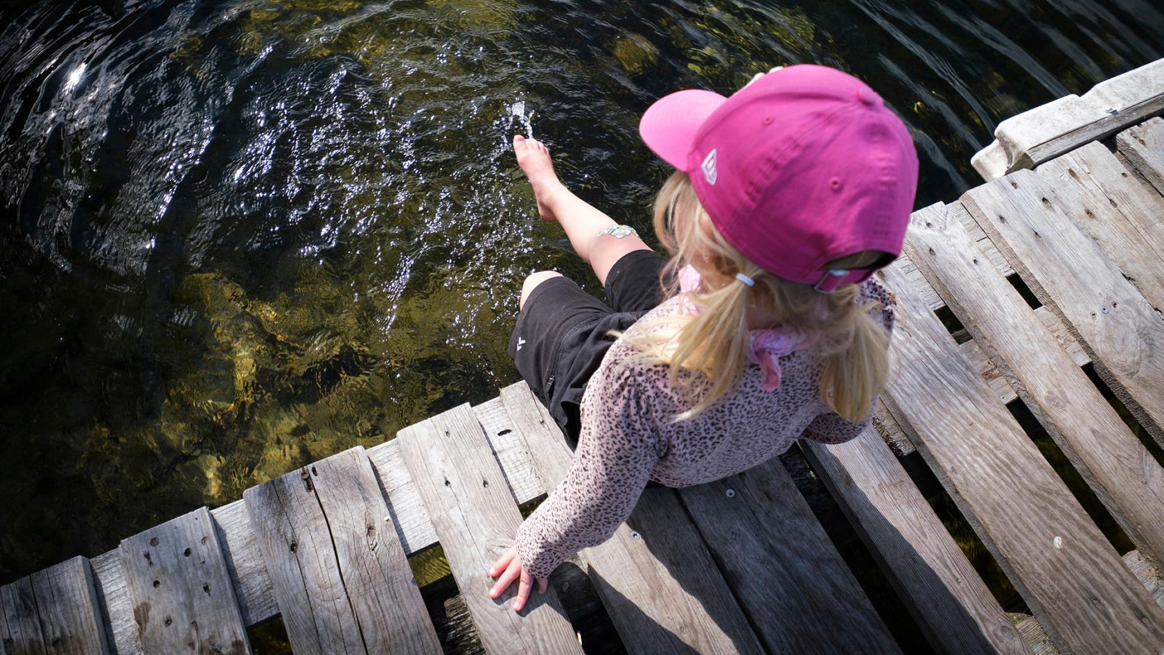 En liten flicka med rosa keps sitter på en brygga och doppar i tårna i vattnet.